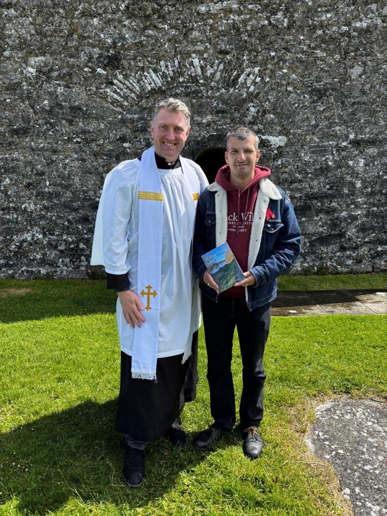 Fr Philip McKinley, wearing white robes and a stole, stands beside Michael McFarland Campbell holding A Living Cloud of Irish Witnesses outside the stone doorway of Temple Connor at Clonmacnoise.